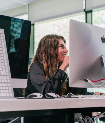 En un aula de UNIE, vemos una alumna sonriente que atiende la clase y trabaja con un mac de pantalla grande.