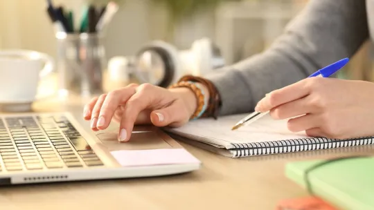 Manos escribiendo en portátil y tomando notas con boli.