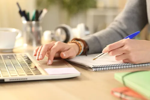 Manos escribiendo en portátil y tomando notas con boli.